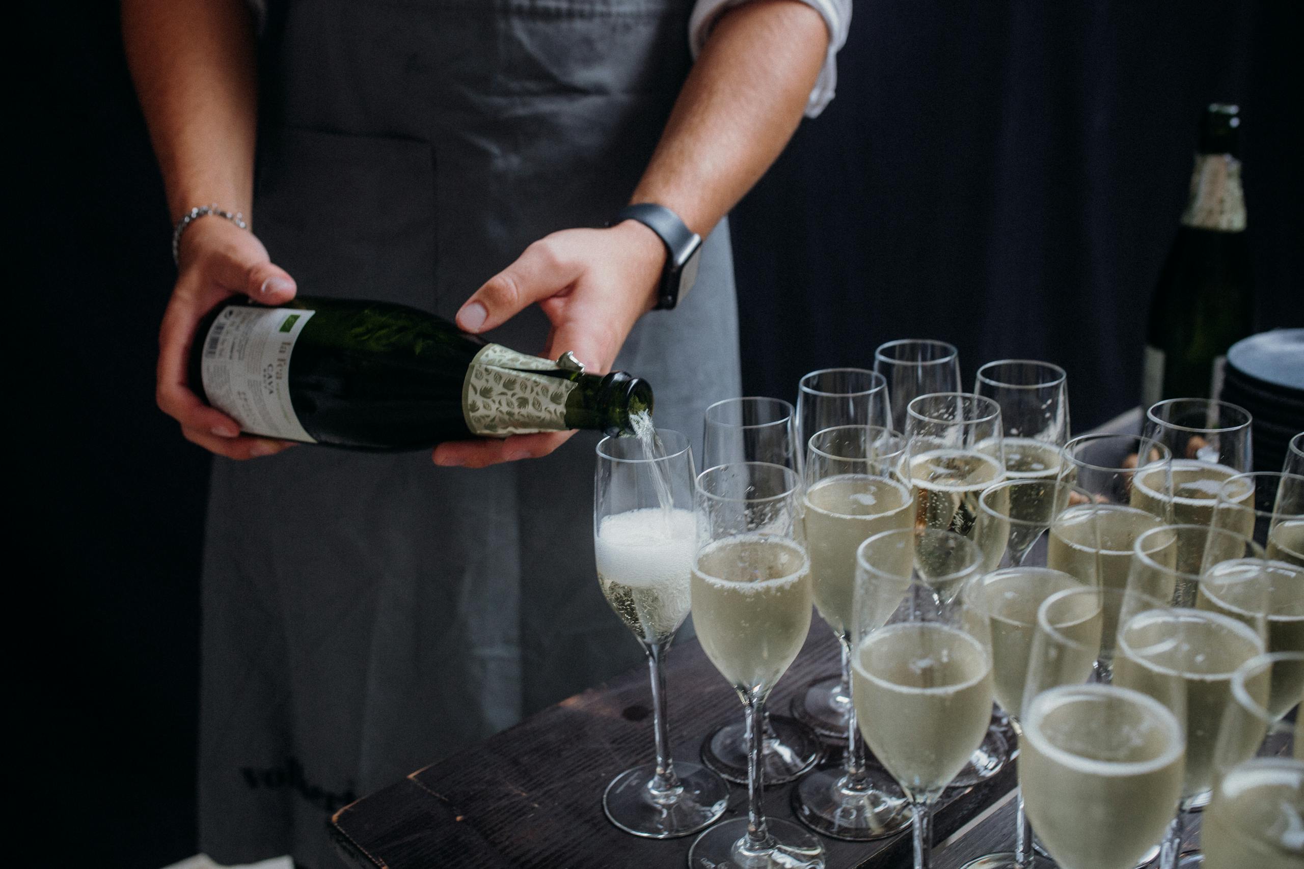 A bartender pouring champagne into multiple glasses on a wooden table at an event.